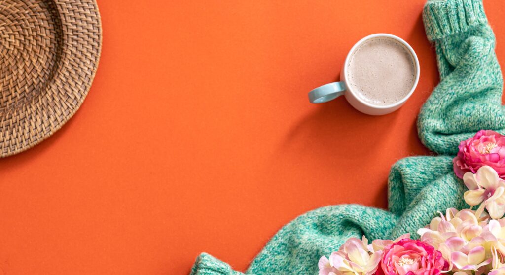 Cup of coffee, flowers and knitted element on orange background, flat lay.